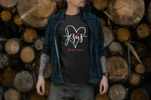 Hombre con camiseta negra que muestra el texto 'Jesus' en caligrafía blanca con un corazón estilizado y 'Es mi Señor' en rojo, combinada con una camisa de cuadros verde. Ideal para regalos cristianos, ropa de fe y expresión religiosa con mensaje inspirador.
